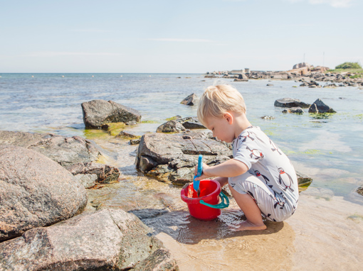Kleiner Junge mit rotem Eimer spielt barfuß auf den flachen Felsen am Sandstrand von Sandvig